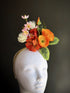 Floral fascinator hat with orange, coral, and pink flowers and green leaves on beige headband

