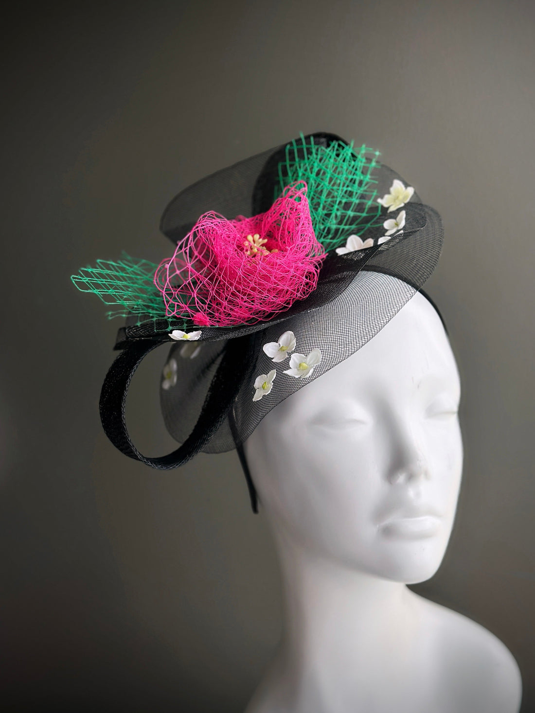 Black mesh fascinator hat with pink netting, green accents, and white flower embellishments