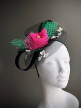 Black mesh fascinator hat with pink netting, green accents, and white flower embellishments