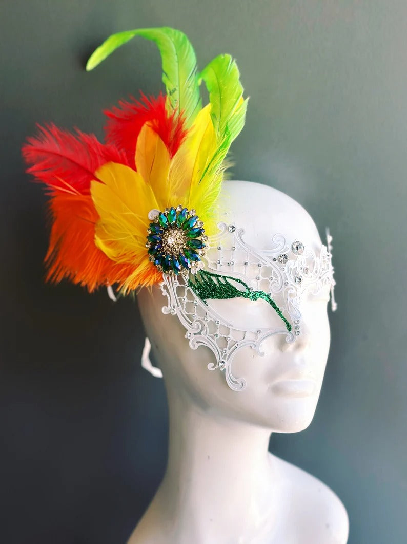 White masquerade mask with filigree design, rhinestones, and red yellow green feathers.