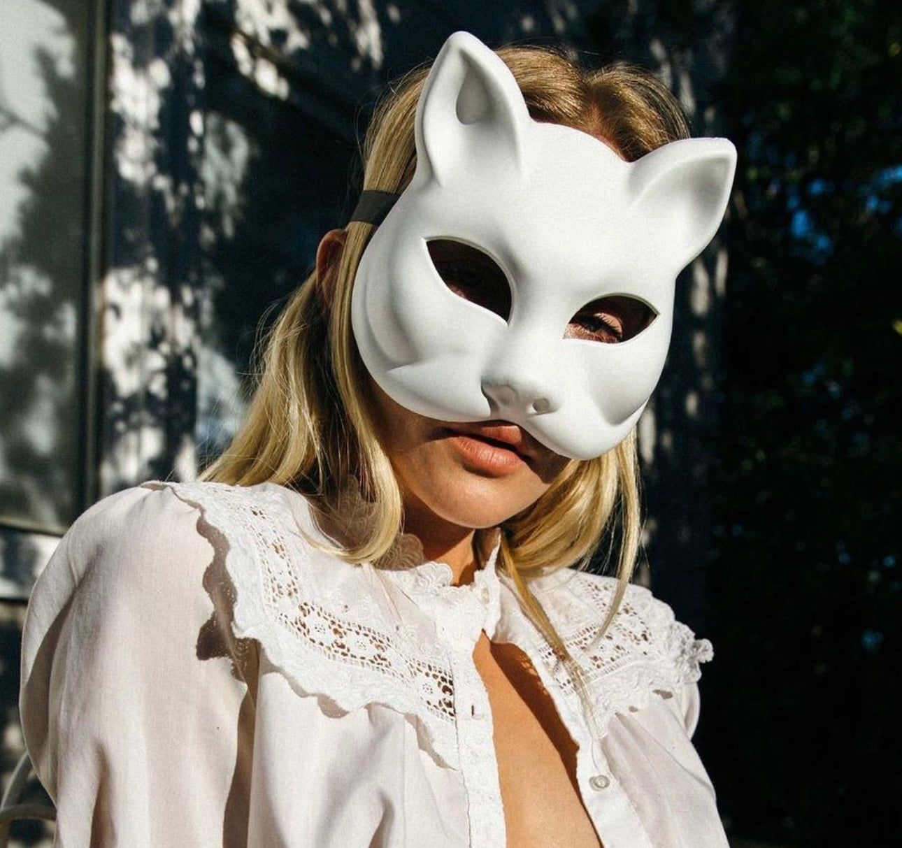 A person wearing a white therian mask with cat-like features, including ear-like protrusions and a nose detail, in a sunlit outdoor setting with a white ruffled garment.